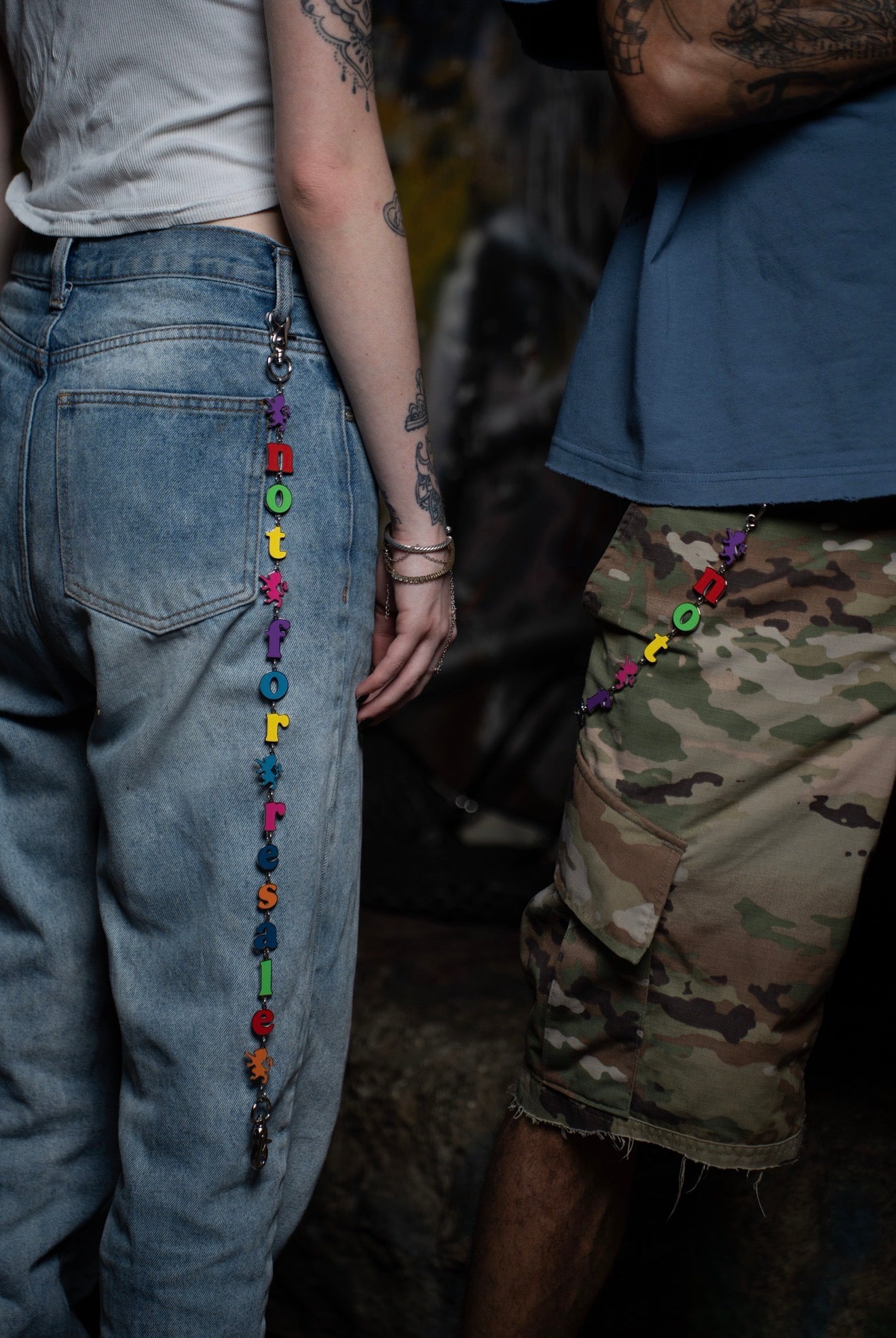 A close-up lifestyle shot of two people standing side-by-side, each wearing a colorful beaded chain accessory draped down the back of their jeans. The person on the left wears light-wash blue jeans, and the person on the right wears camouflage cargo shorts.